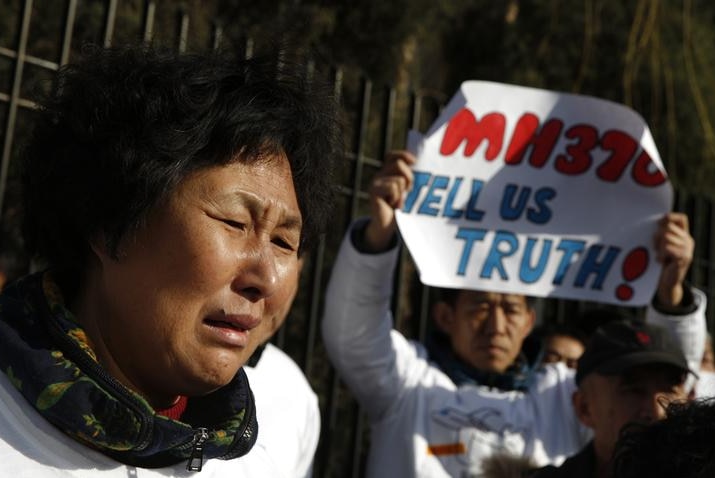 Liu Guiqiu is seen to the left of the shot crying, as a man stands in the background with sign that says "MH370 TELL US TRUTH".