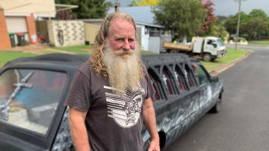 A man with long hair and a long grey beard stands in front of a gothic-looking hearse vehicle