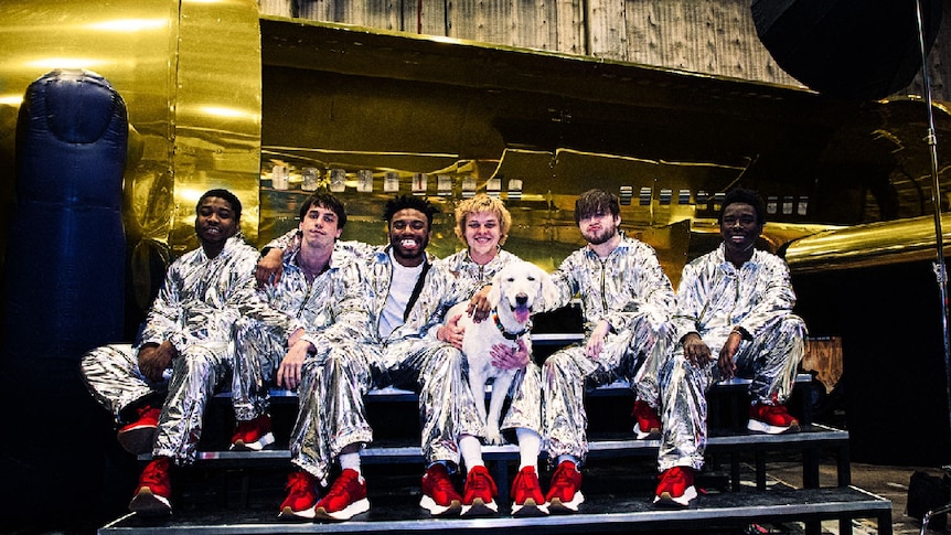 Image of BROCKHAMPTON dressed in silver suits and red shoes, sitting with a dog