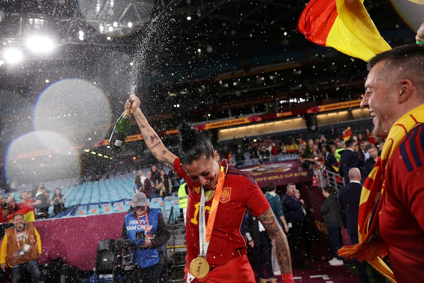 A woman wearing a red football uniform and a gold metal around her neck lifts a bottle of spraying champagne