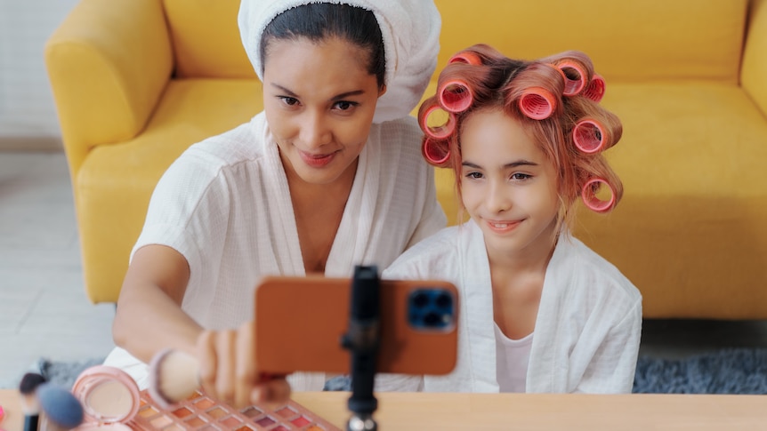 Mother and daughter doing hair and makeup in front of smartphone, filming themselves. 