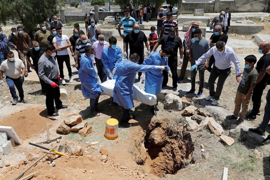 Mourners wearing masks stand around four men holding a body as it is lowered into a dug up grave