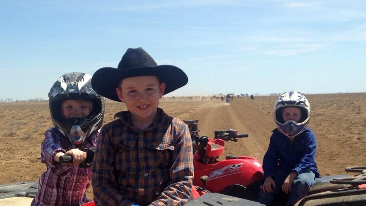 Three young children sit on quad bikes in the outback.