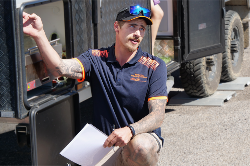 A young man wearing a cap kneels beside the front of a caravan, looking up to explain something to the driver