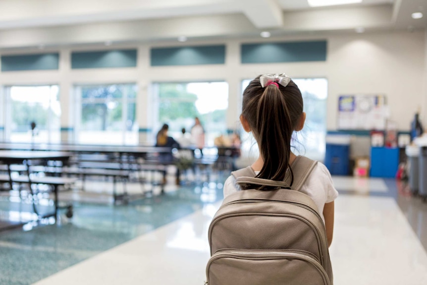 Anonymous photo of a young girl wearing a back pack in a school lunch room.