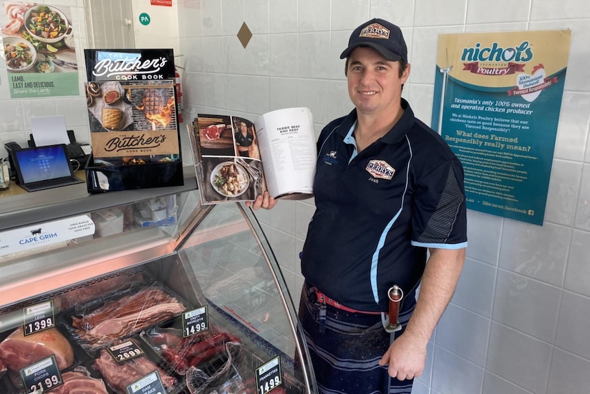 Josh Perry stands next to a a meat display in his butcher shop holding a recipe book