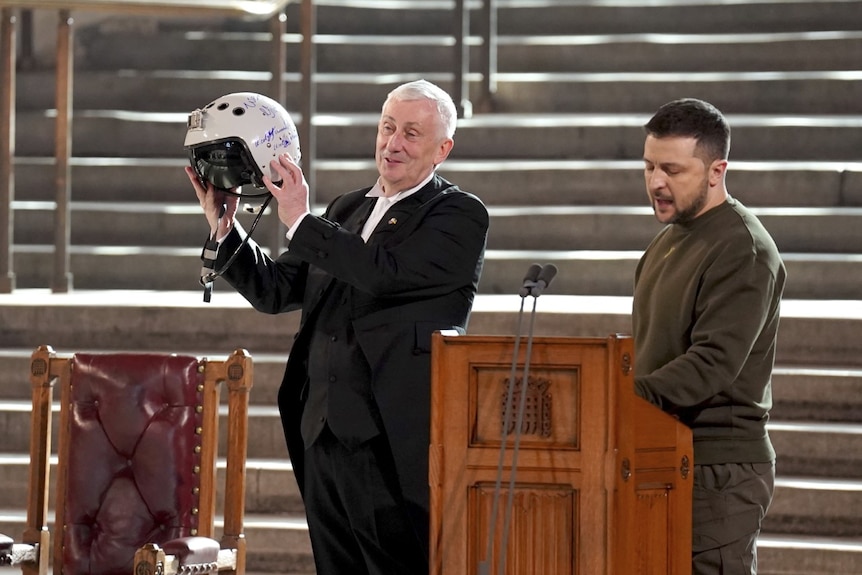 Zelenskyy stands at a wooden lectern. On the left, a man in a suit holds up a white helmet decorated with handwriting