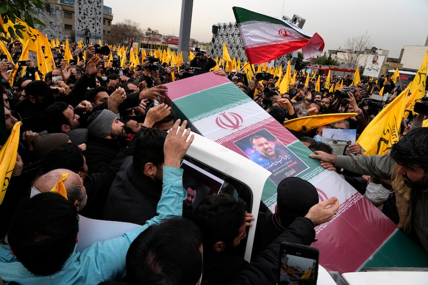 Mourners carry the flag-draped coffin of a high ranking general at a funeral with large crowd, many carrying yellow flags.