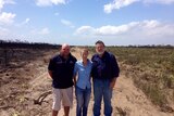 Two men and one woman stand on bare earth between burnt bush and green bushland