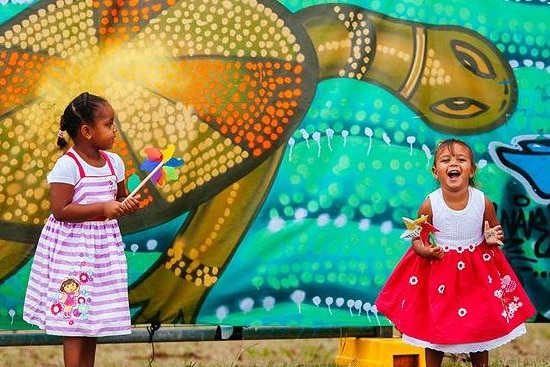 Two young Indigenous girls standing in front of a wall with a mural painted on it.