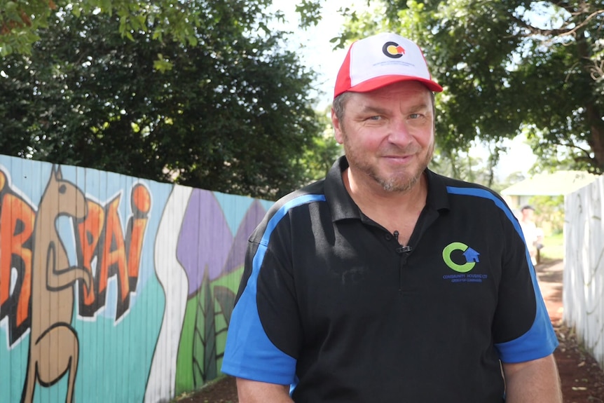 Man in black and blue t-shirt stands in front of mural featuring kangaroo and word 'Birpai' 