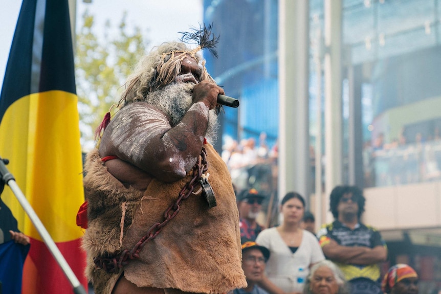 An Aboriginal man in traditional dress speaks into a microphone at a rally