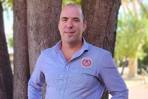 A man smiles at the camera, standing in front of a tree wearing a blue button down shirt.