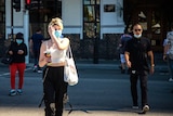 A woman wearing a mask and holding a drink crosses a city street.