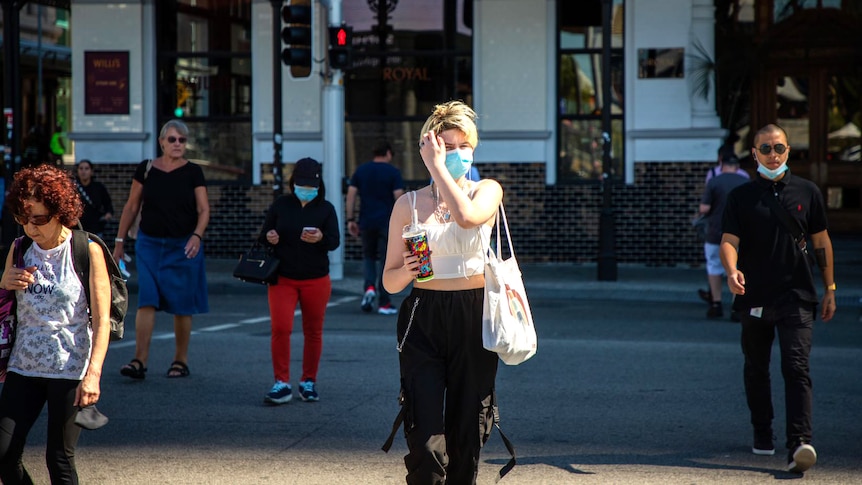 A woman wearing a mask and holding a drink crosses a city street.
