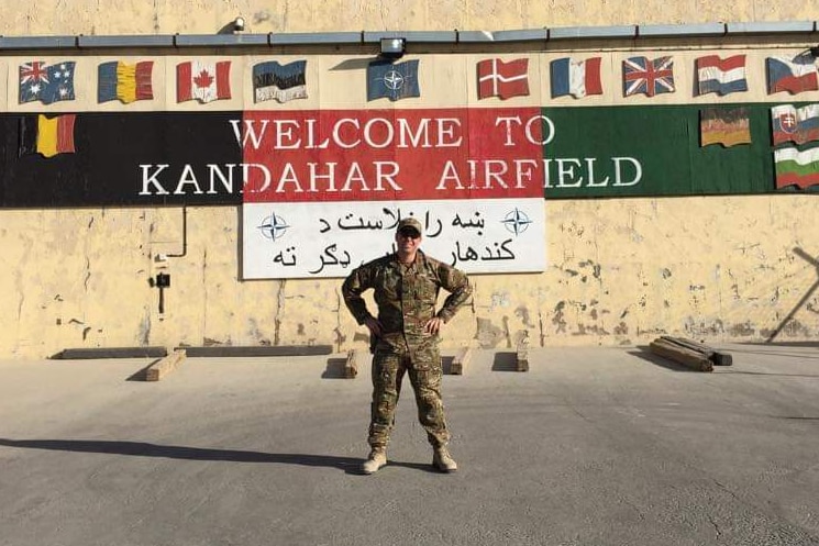 A man wearing a military uniform stands in front of a hangar.