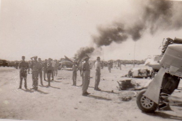 An old sepia tones image of soldiers with smoke billowing from a crashed plane in the background.