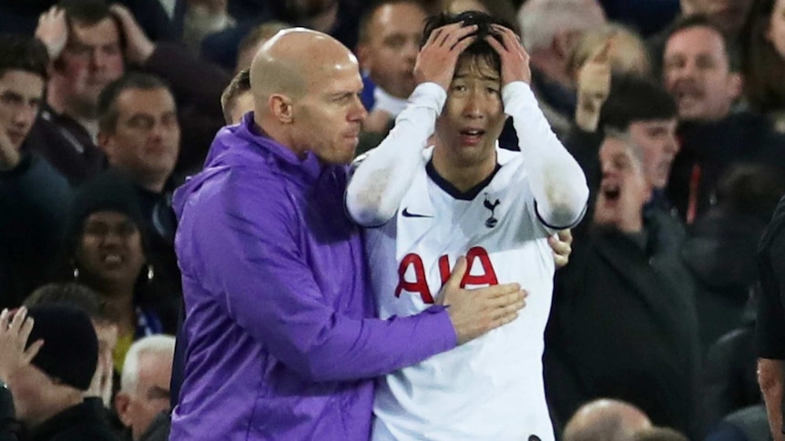 Son Heung-Min holds his head in his hands as a man in a blue jacket hugs him and pats his chest