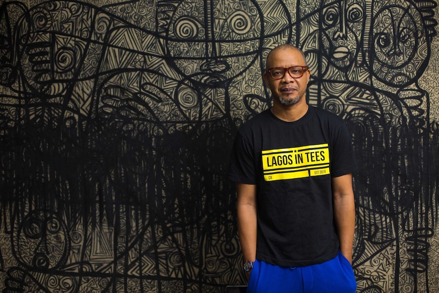 Victor stands in front of a large artwork of figures with complicated patterns drawn in black on a white background.