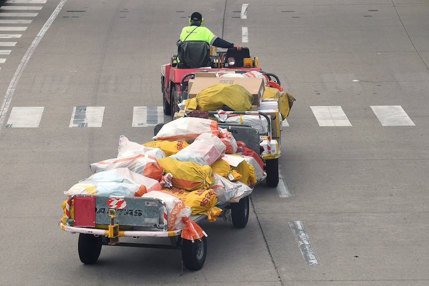 A Qantas worker drives a cart with a trailer behind loaded with parcels on a tarmac.