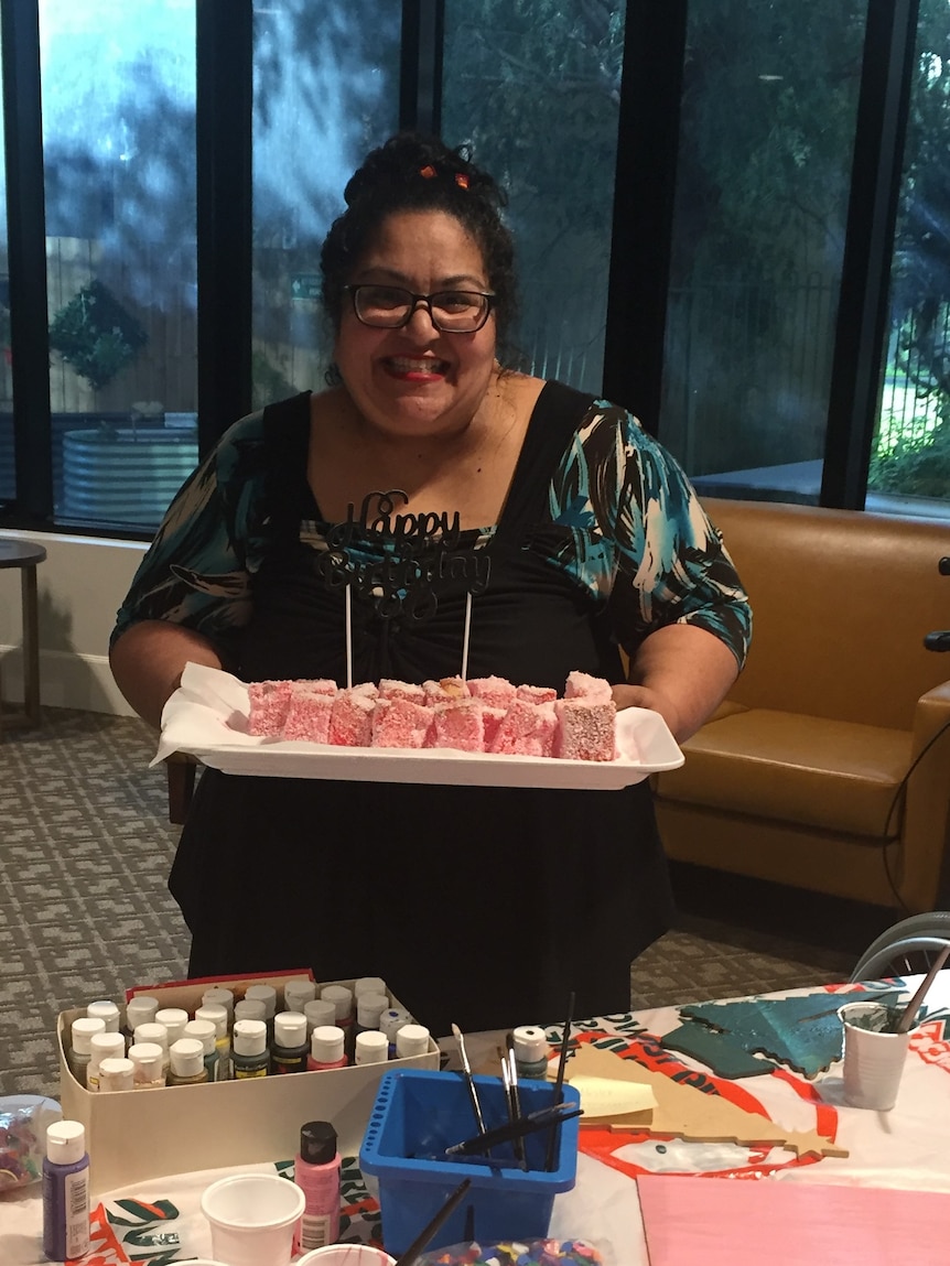 Aged care worker Sherene Magana-Cruz smiling and standing in front of a table holding a cake at work 