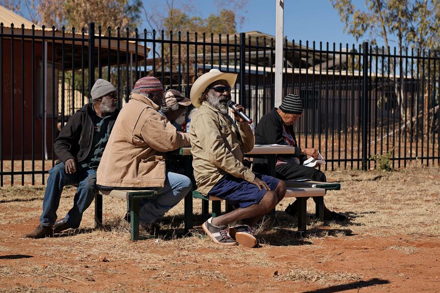 Otto sims talks into the microphone sitting with community members at a table in Yuendumu.