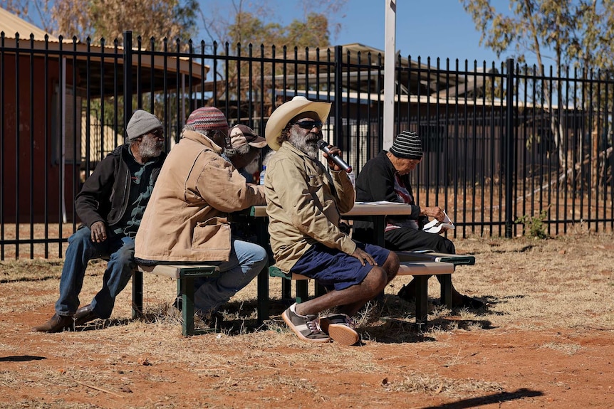 Otto sims talks into the microphone sitting with community members at a table in Yuendumu.