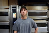 Man wearing cap and grey t-shirt standing in front of electric ovens