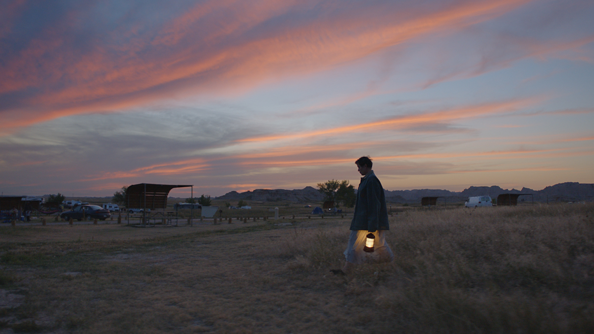 Sunset shot of the actress with pixie haircut wearing coat and carrying hurricane lamp, walking across grass in countryside.