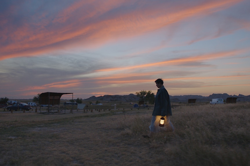 Sunset shot of the actress with pixie haircut wearing coat and carrying hurricane lamp, walking across grass in countryside.