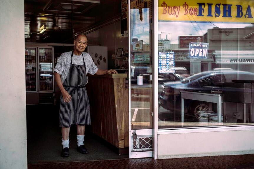 72-year-old  man stands in the doorway of an old-fashioned fish and chip shop.