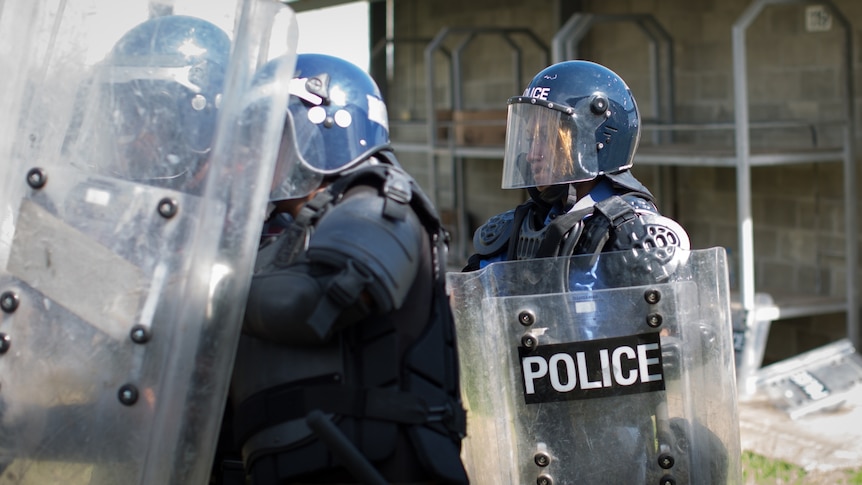 A police officer wearing a helmet and protective equipment, and carrying a large plastic shield, receives training
