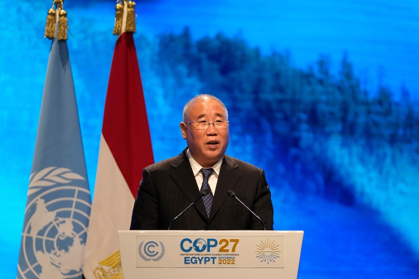 A man in a suit speaks from behind a lecturn with two flags hanging behind.