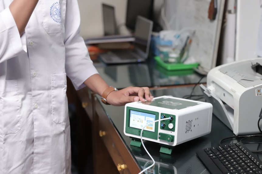 A woman points to a device with a screen and a cord plugged in.