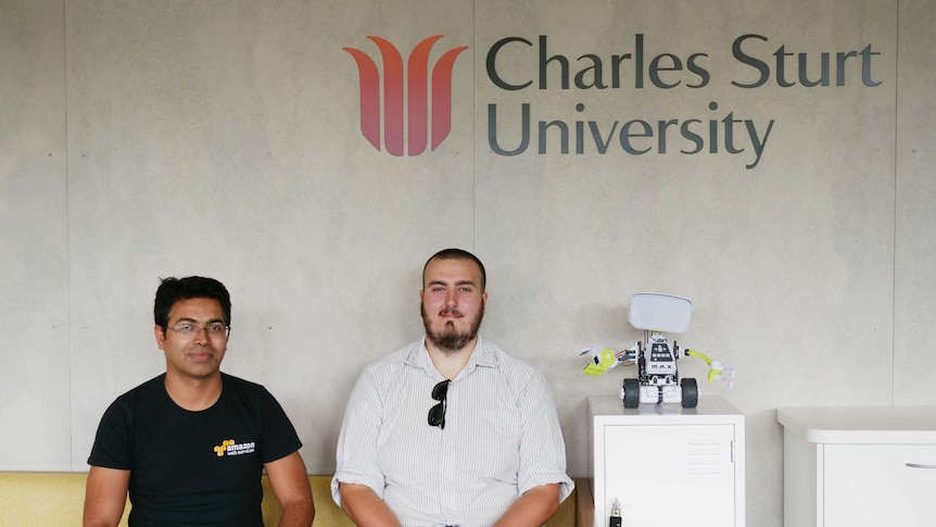 Two men sitting together on a couch below a sign that says Charles Sturt University