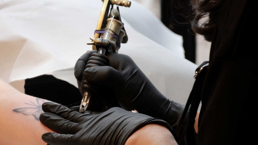 A woman holds a tattoo gun to a client's arm.