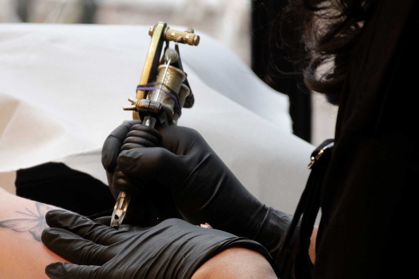 A woman holds a tattoo gun to a client's arm.