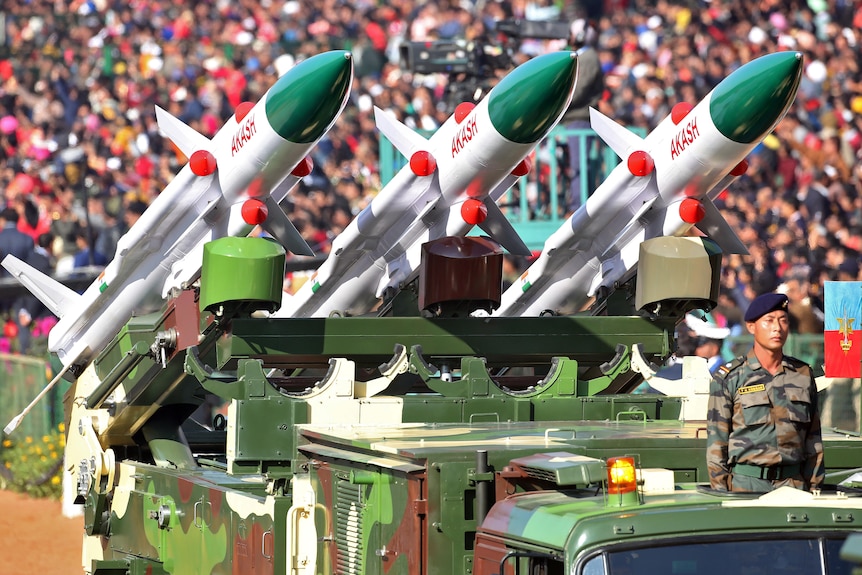 A line of three missiles with Akash written on the side sit on a truck in front of a crowd.