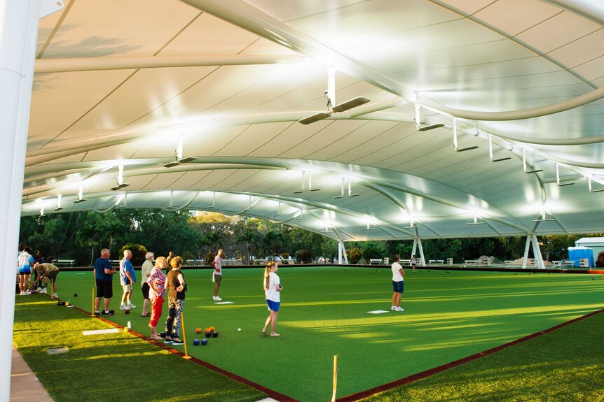People playing lawn bowls on an artificial lawn under a large white shade. Evening light. Artificial lights on.