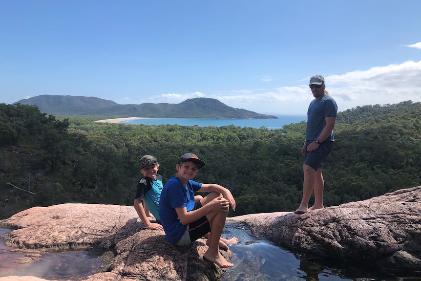A man and two boys sit on rocks at the top of a mountain overlooking a beach and the sea with rainforest all around.