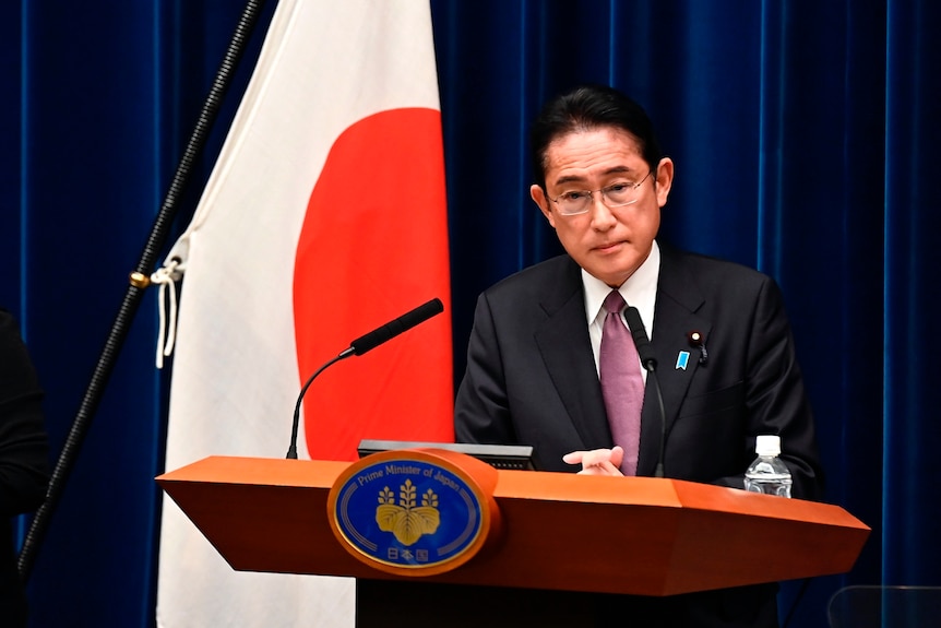 Fumio Kishida attends a press conference and speaks behind a podium on stage, next to a Japanese flag.