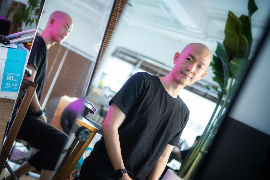 A bald Korean man in a black t-shirt sits on a chair with his reflection in a mirror behind him 