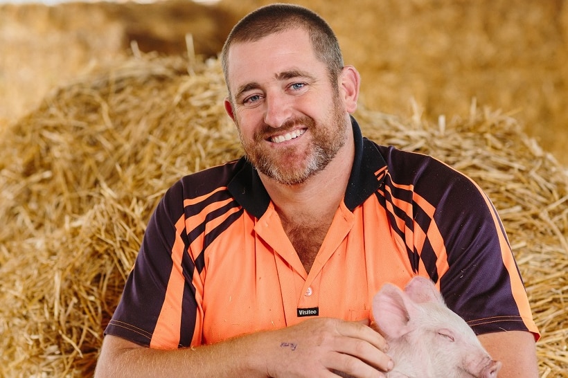 A man sitting on hay bales, smiling and holding a piglet.