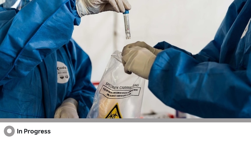 A man wearing full PPE holds a specimen bag while another man wearing full PPE drops a specimen tube inside