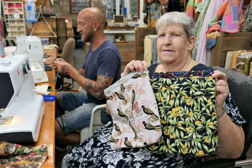 Older woman, holding up fabric pouches, and younger man sit at sewing machines