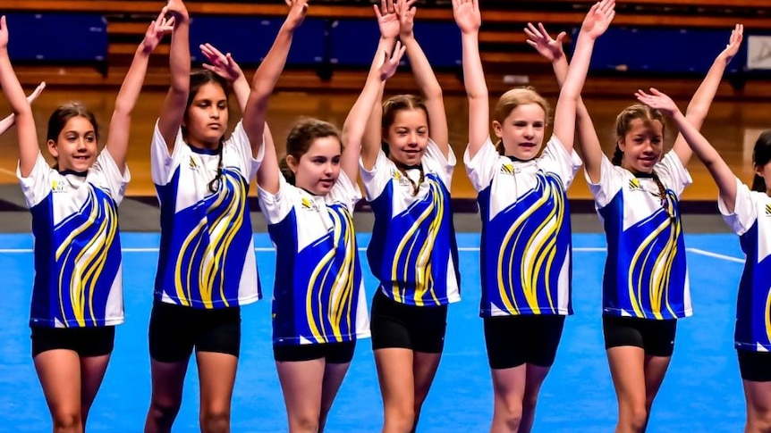 seven young gymnasts wearingwhite and blue tops and black shorts with hands raised standing on blue mats