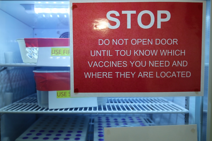 A red laminated sign on a small fridge reads 'STOP: DO NOT OPEN DOOR UNTIL YOU KNOW WHICH VACCINES YOU NEED'