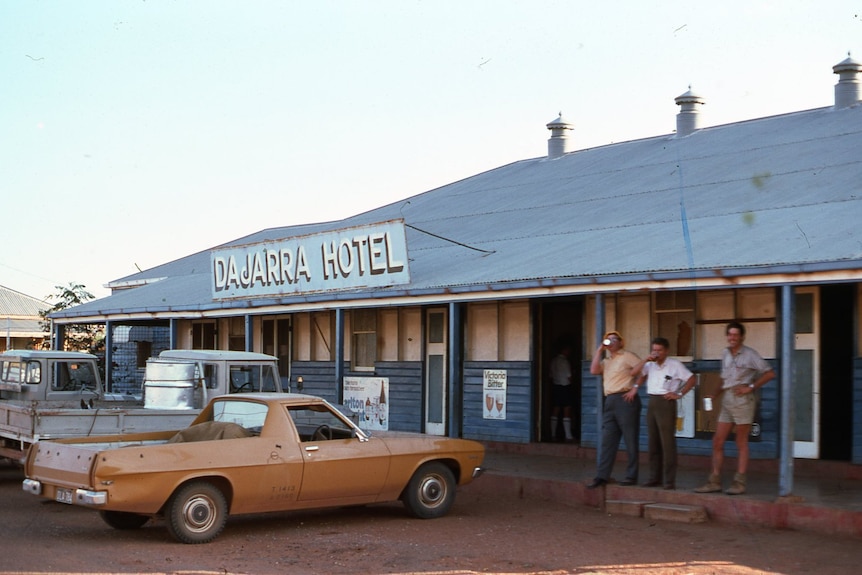 A squat outback pub with several utes parked out the front and a group of men knocking back drinks on the veranda.