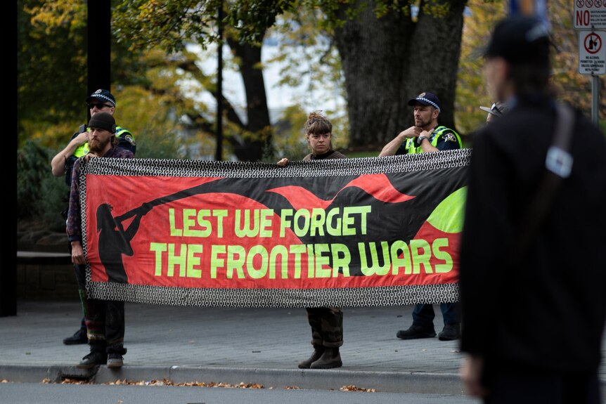 Three people hold a banner that reads "Lest we forget the frontier wars" on a city street