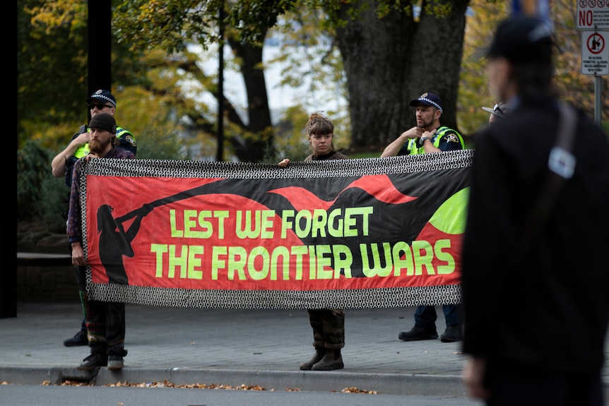 Three people hold a banner that reads "Lest we forget the frontier wars" on a city street